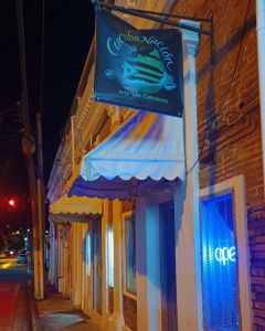 Brick store front scene at night with white awning, blue flourescent sign that reads OPEN glowing and a blue and green banner on a black background that reads CucubaNacion.